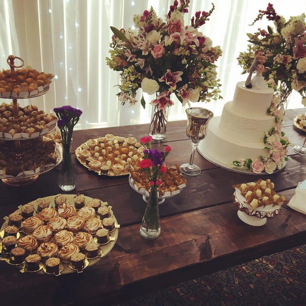 Wedding dessert table with a white tiered cake, flowers and trays of Brazilian sweets